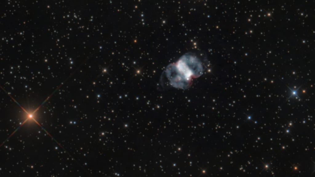 Astrophotography image of the Little Dumbell Nebula. A thick white bar flanked by blue loops on a field of stars.