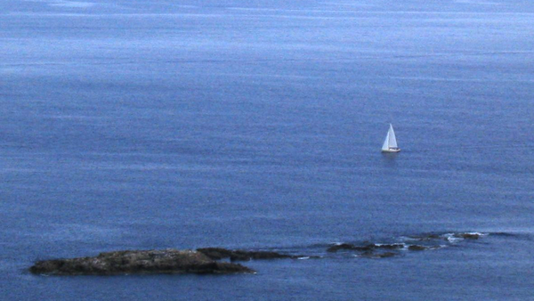 A small sailboat passes behind a foreground rock reef in calm blue water 
